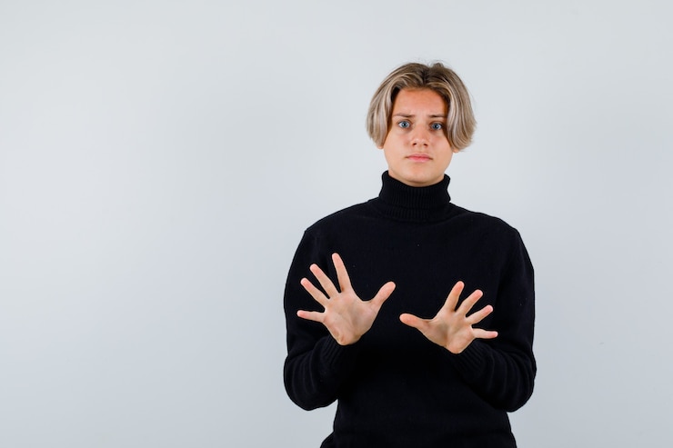 Foto grátis retrato de um lindo menino adolescente mostrando um gesto de rendição com uma blusa de gola alta preta e olhando a vista frontal com medo