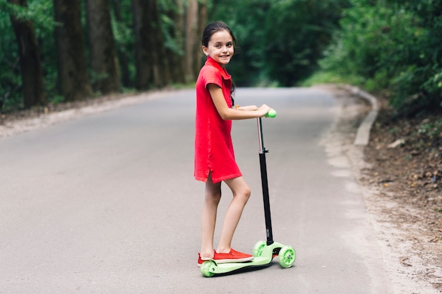 Retrato, de, um, menina sorridente, ficar, sobre, empurre scooter, ligado, estrada