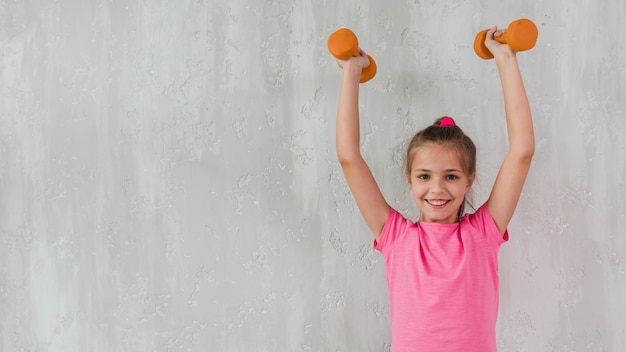 Retrato, de, um, menina sorridente, levantamento, dela, mãos, segurando, dumbbells, frente, parede concreta