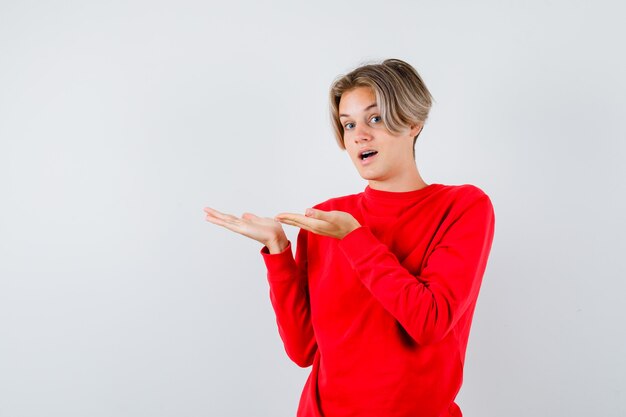 Retrato de um menino adolescente fingindo mostrar algo, abrindo a boca em um suéter vermelho e olhando a vista frontal pasmo