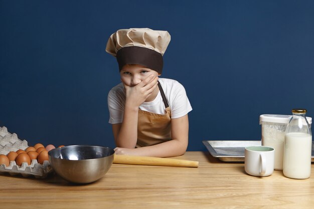 Retrato de um menino de 10 anos frustrado com uniforme de chef cobrindo a boca e sentindo-se confuso enquanto vai fazer panquecas sozinho pela primeira vez com leite