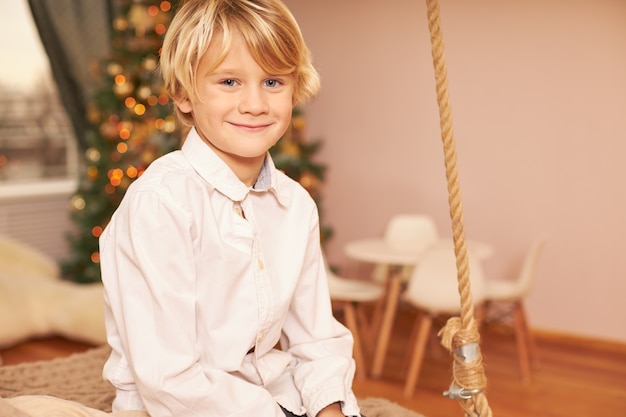 Retrato de um menino europeu fofo vestindo uma camisa branca, curtindo o clima festivo, antecipando a véspera de natal, sentado na sala de estar com a árvore de ano novo decorada, sorrindo feliz