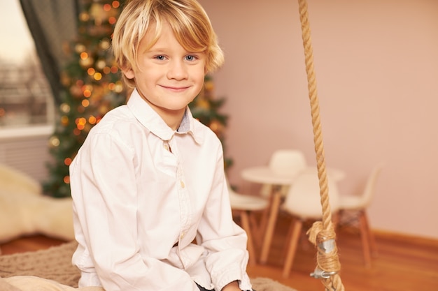 Retrato de um menino europeu fofo vestindo uma camisa branca, curtindo o clima festivo, antecipando a véspera de Natal, sentado na sala de estar com a árvore de ano novo decorada, sorrindo feliz