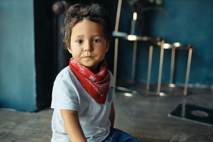 Foto grátis retrato de um menino fofo gordinho mestiço com cabelo encaracolado e pele escura sentado no chão em casa usando um pano vermelho no pescoço, com uma expressão facial triste