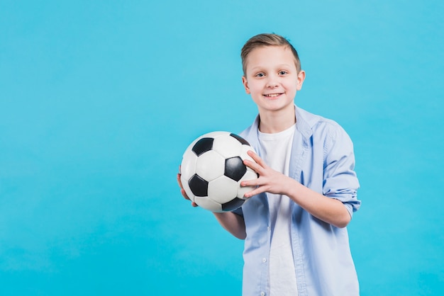 Foto grátis retrato, de, um, menino sorridente, segurando, bola futebol, em, mão, ficar, contra, céu azul