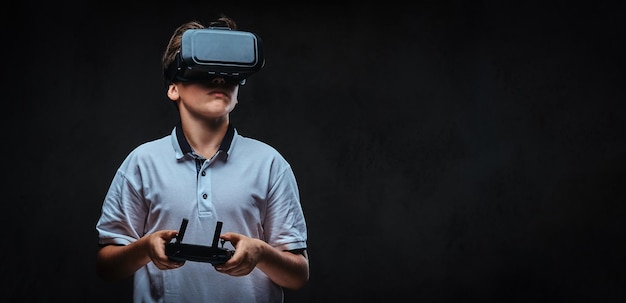 Retrato de um menino vestido com uma camiseta branca usando óculos de realidade virtual e detém um controle remoto. Isolado no fundo escuro.