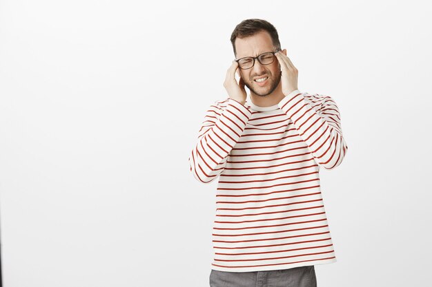 Retrato de um modelo desconfortável de homem sofrendo de óculos, fechando os olhos e fazendo careta de dor, segurando as têmporas de mãos dadas