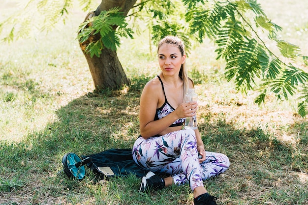 Foto grátis retrato, de, um, mulher jovem, sentando, perto, a, árvore, segurando, garrafa água, em, mão