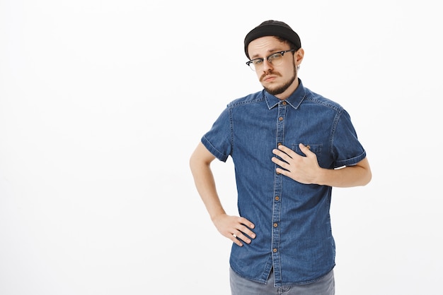 Retrato de um músico habilidoso, mandão, confiante e bonito, com um gorro preto da moda e uma camisa apontando para si mesmo com a palma da mão no peito apertando os olhos e olhando intrigado e duvidoso