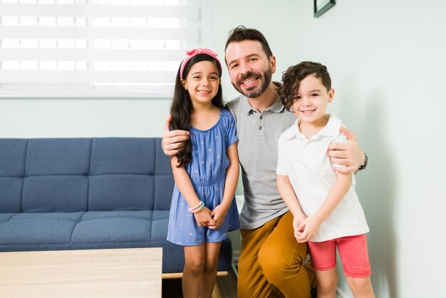 Retrato de um pai bonito abraçando seu menino e menina enquanto sorria enquanto fazia contato visual em casa. Pai latino tirando uma foto com seus filhos