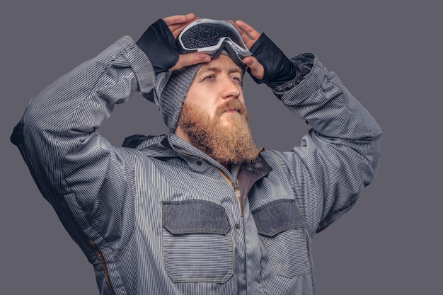 Retrato de um snowboarder ruiva com uma barba cheia em um chapéu de inverno e óculos de proteção vestido com um casaco de snowboard posando em um estúdio, olhando para longe. Isolado no fundo cinza.