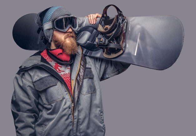 Retrato de um snowboarder vestido com um equipamento de proteção completo para snowboard extream posando com uma prancha de snowboard no ombro em um estúdio. Isolado em um fundo cinza.