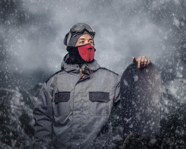 Retrato de um snowboarder vestido com um equipamento de proteção completo para snowboard extremo posando com snowboard no contexto das montanhas