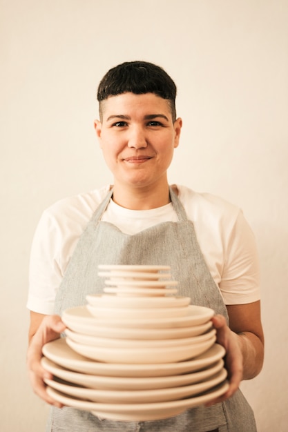 Foto grátis retrato, de, um, sorrindo, femininas, oleiro, segurando, pilha, de, dishware, em, mão, contra, bege, fundo