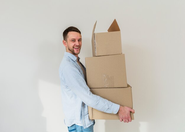 Retrato, de, um, sorrindo, homem jovem, carregar, pilha, de, caixas cartão, contra, parede branca