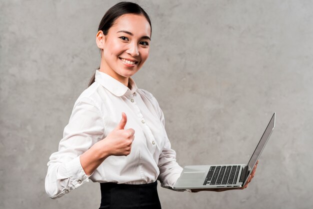 Retrato, de, um, sorrindo, jovem, executiva, segurando, laptop, em, mão, mostrando, polegar, sinal