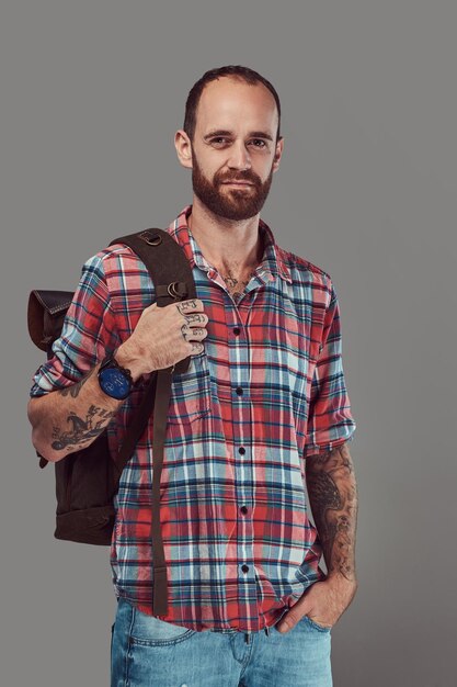 Retrato de um viajante tatuado bonito em uma camisa de flanela com uma mochila, de pé em um estúdio. Isolado em um fundo cinza.