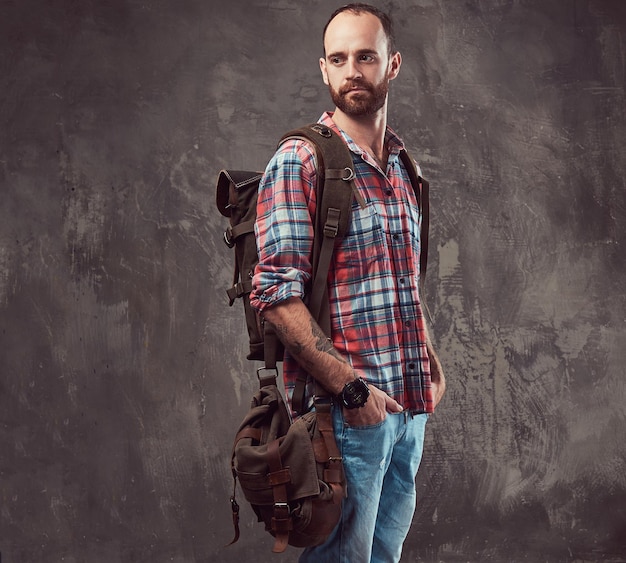 Retrato de um viajante tatuado bonito em uma camisa de flanela com uma mochila, de pé em um estúdio. Isolado em um fundo cinza.