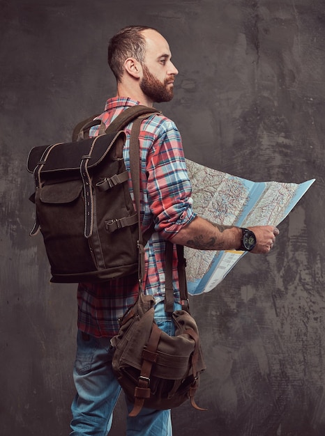 Retrato de um viajante tatuado bonito em uma camisa de flanela com uma mochila, segura um mapa, de pé em um estúdio. Isolado em um fundo cinza.