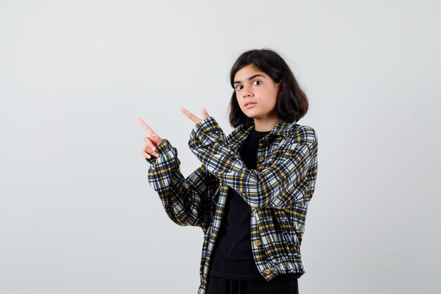 Retrato de uma adolescente apontando para o canto superior esquerdo com uma camisa casual e olhando para a frente em choque