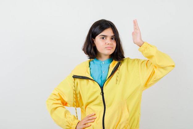 Retrato de uma adolescente levantando a mão, segurando a mão na cintura com uma jaqueta amarela e olhando de frente com confiança