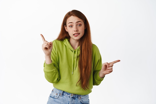 Retrato de uma adolescente ruiva indecisa encolhendo os ombros e apontando para duas maneiras, os lados esquerdo e direito parecem confusos confusos sobre o que escolher fundo branco