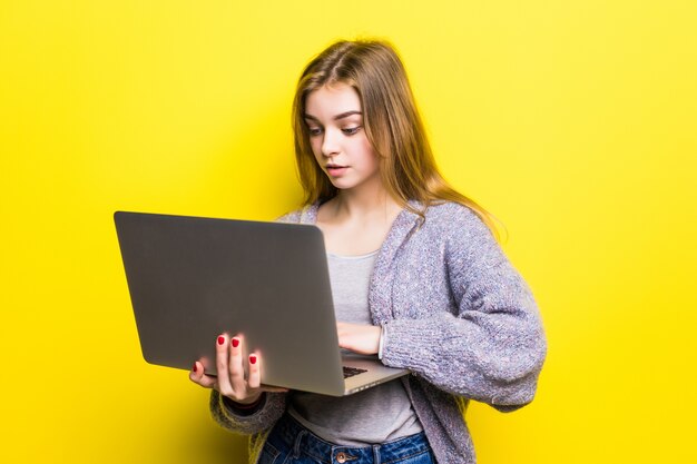 Retrato de uma adolescente sorridente segurando um laptop isolado em uma parede amarela