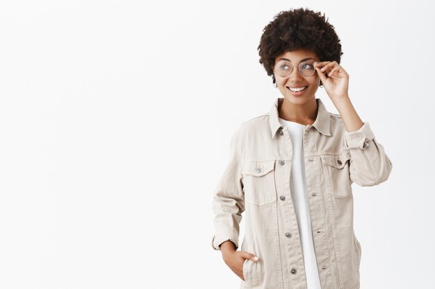 Retrato de uma adorável mulher adulta afro-americana com uma camisa bege e óculos tocando a borda dos óculos enquanto sorri alegremente e olhando para a esquerda para verificar as lojas que passavam pela cidade
