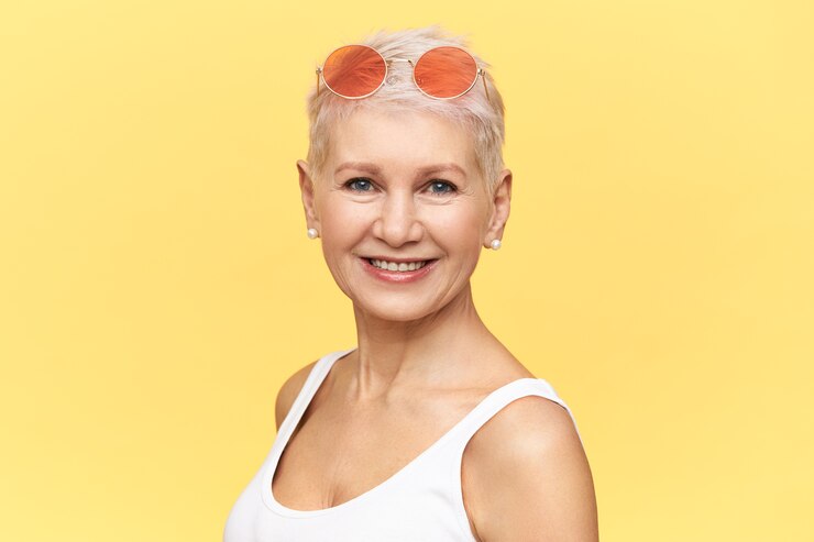 Foto grátis retrato de uma aposentada caucasiana na moda, caminhando em um dia ensolarado e quente, usando óculos de sol redondos rosa na cabeça, olhando para a câmera com um sorriso feliz