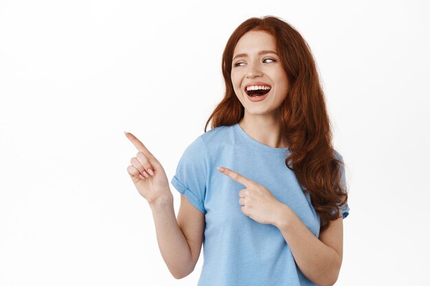 Retrato de uma autêntica garota ruiva feliz rindo, sorrindo e apontando para a esquerda com uma expressão de rosto alegre, de pé no branco.