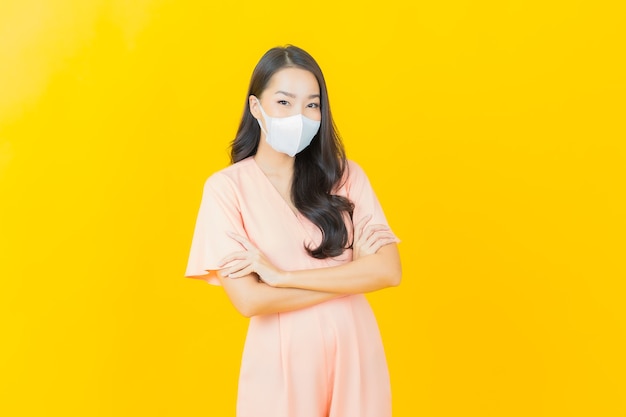 Retrato de uma bela jovem asiática com máscara para proteger o vírus covid19 na parede de cor amarela