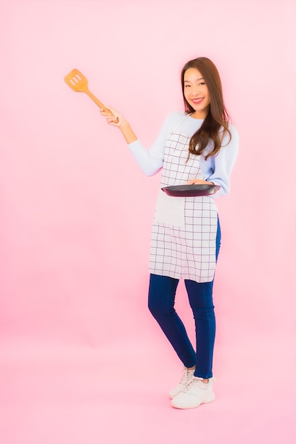 Retrato de uma bela jovem asiática em roupas de cozinha com avental na parede rosa isolada