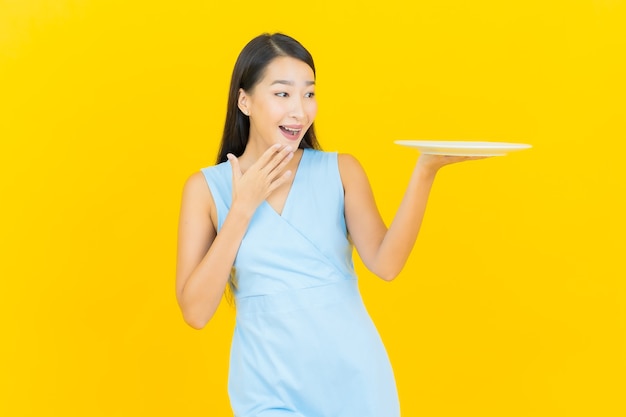 Retrato de uma bela jovem asiática sorrindo com prato vazio na parede de cor amarela.