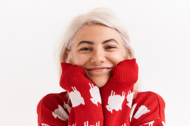 Retrato de uma bela jovem europeia com penteado bob bagunçado e piercing facial, posando na parede branca em um jumper vermelho elegante, colocando o queixo nas mangas esticadas e sorrindo