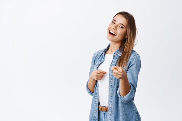 Retrato de uma bela jovem loira em roupa casual, sorrindo e apontando o dedo, elogiando ou convidando vocês, em pé no branco