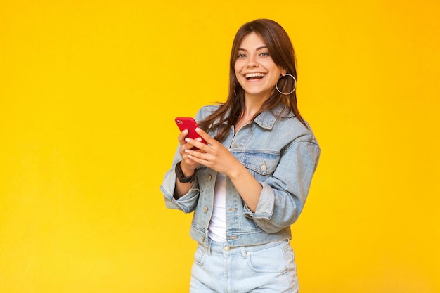 Retrato de uma bela jovem morena feliz com maquiagem em jeans estilo casual em pé, segurando seu telefone móvel inteligente vermelho, olhando com um sorriso. tiro do estúdio, isolado no fundo amarelo.