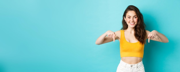 Foto grátis retrato de uma bela jovem mostrando a direção apontando os dedos para baixo no anúncio sorrindo alegre recomendando check-out em pé sobre fundo azul