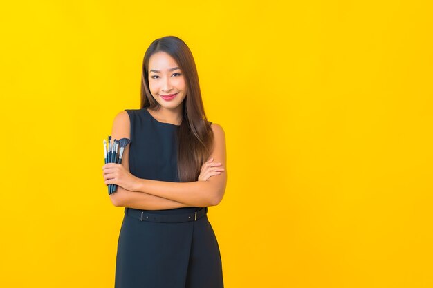 Retrato de uma bela jovem mulher de negócios asiática com escova cosmética de maquiagem em fundo amarelo.