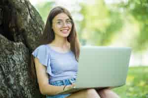 Foto grátis retrato de uma bela jovem sentada na grama verde no parque com as pernas cruzadas durante o dia de verão, usando um laptop