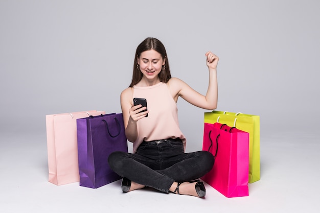 Retrato de uma bela jovem sentada no chão com sacolas de compras e usando telefone celular com gesto de vitória sobre parede cinza