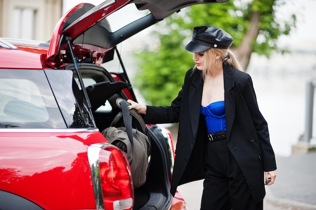 Retrato de uma bela loira sexy modelo de mulher de boné e todo preto com maquiagem brilhante perto do carro vermelho da cidade colocou sua bolsa no porta-malas