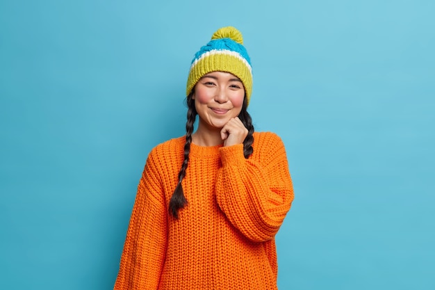 Retrato de uma bela morena tímida mulher asiática com duas tranças usa um suéter de malha e um chapéu satisfez as poses de expressão facial no estúdio contra a parede azul caminha durante o inverno