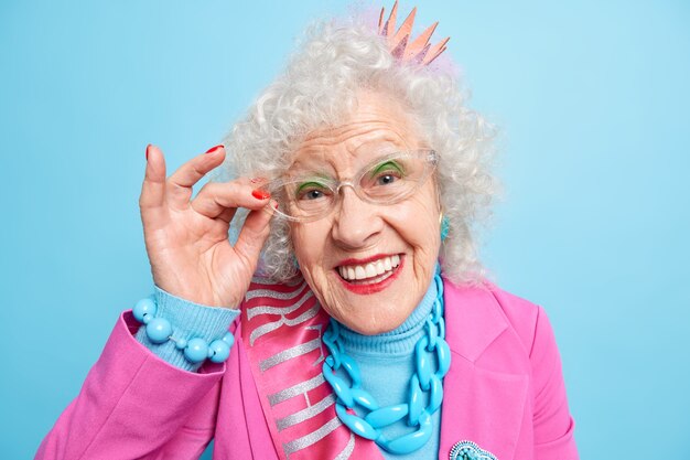 Retrato de uma bela mulher idosa de cabelos grisalhos segurando a mão na borda dos óculos e sorrindo alegremente