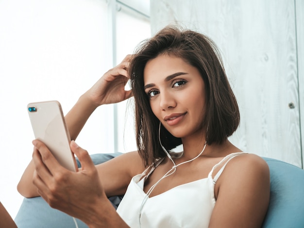 Foto grátis retrato de uma bela mulher sorridente, vestida de pijama branco e ouvindo música com fones de ouvido. modelo despreocupado sentado na cadeira macia.