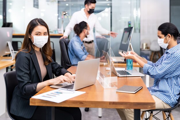 Retrato de uma empresária asiática de funcionário de escritório usa máscara protetora para trabalhar no novo escritório normal com a equipe interracial em segundo plano, pois a prática de distância social evita