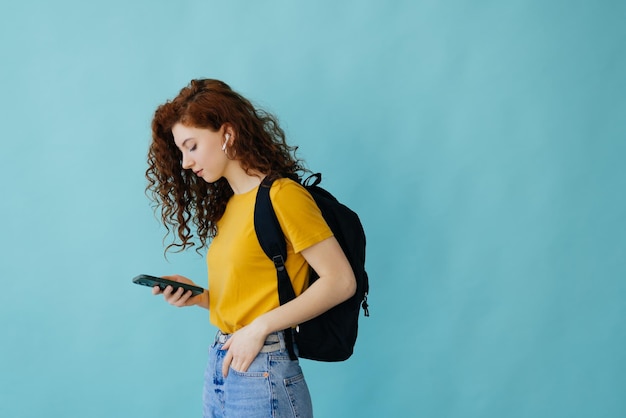 Retrato de uma estudante adorável com mochila ouvindo música com fones de ouvido em pé e segurando o celular isolado sobre fundo azul