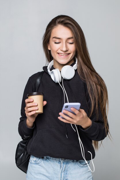 Retrato de uma estudante atraente alegre com mochila, ouvindo música com fones de ouvido enquanto mostra o telefone móvel de tela em branco e dança isolado sobre a parede branca