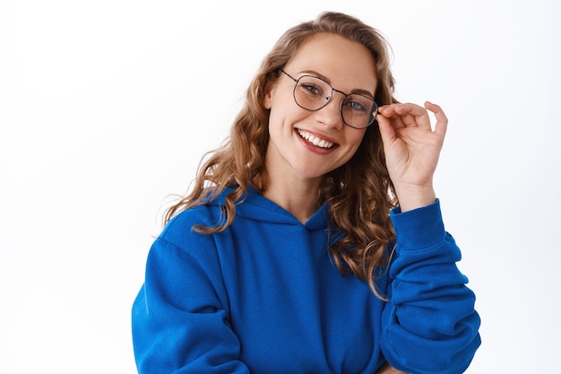 Retrato de uma estudante loira atraente, usando óculos e sorrindo determinado, parecendo elegante, em pé sobre uma parede branca