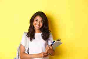 Foto grátis retrato de uma estudante universitária afro-americana feliz, segurando cadernos e uma mochila, sorrindo e em pé sobre um fundo amarelo