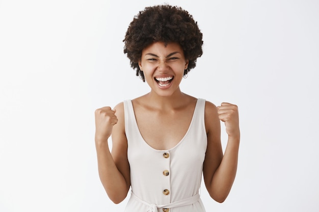 Retrato de uma feliz e satisfeita mulher bonita de pele escura, de macacão branco, levantando os punhos em gesto de vitória e gritando com triunfo